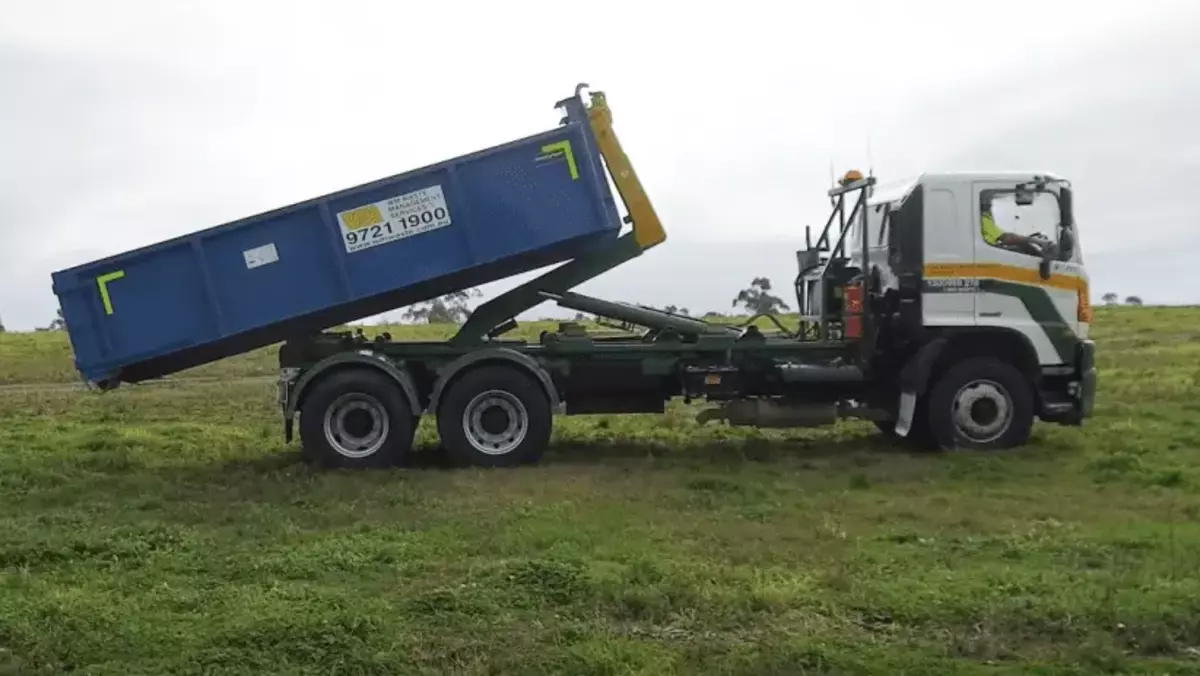 Hook-lift truck tipping a blue bin in a grassy area